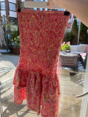 Rosa mönstrad kort klänning - Supersöt kort klänning i rosa och gult med smock upptill och volang nedtill. Klänningen har ett färgglatt mönster och är perfekt för varma dagar. Materialet känns lätt och luftigt, troligen bomull eller polyester. Ingen synlig logga eller märke.