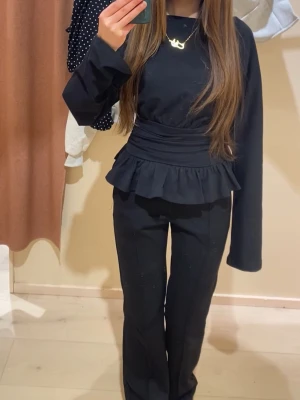 Svart peplum topp med volang - Snygg svart topp med peplum och bred volang i midjan. Långärmad modell med rund halsringning och markerad midja. Perfekt att styla med både jeans och kostymbyxor för en trendig look.