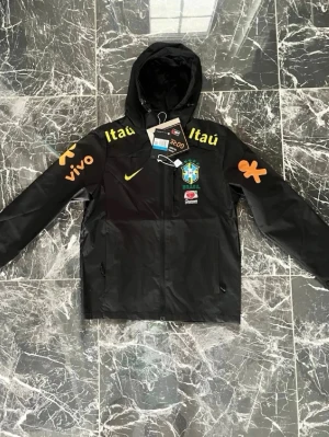 Svart Nike Brasil vindjacka med huva - Snygg svart vindjacka från Nike med Brasiliens landslagslogga på bröstet och gula detaljer. Jackan har huva, dragkedja framtill och tryckta loggor som Itau, Vivo och Guaraná Antarctica på bröst och ärmar. Perfekt för dig som gillar sportig stil.                                            🔴🔴🔴🔴LITEN I STORLEK PASSAR S-M🔴🔴🔴🔴
