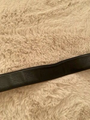 Svart skärp från Gucci i skinn - Säljer ett svart skärp från Gucci i äkta skinn med det klassiska GG-mönstret präglat över hela bandet. Bältet har en bred design och känns riktigt lyxigt. Perfekt för dig som vill addera en exklusiv touch till din outfit.