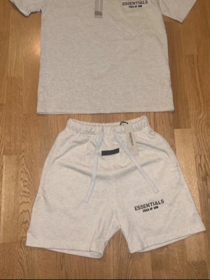 Essentials loungewear set i grått - Säljer ett loungewear set från Fear of God Essentials i ljusgrått. Setet består av en t-shirt med rund hals och tryck samt matchande shorts med dragsko och elastisk midja. Båda plaggen har Essentials-logga tryckt framtill. Perfekt för chill dagar hemma.