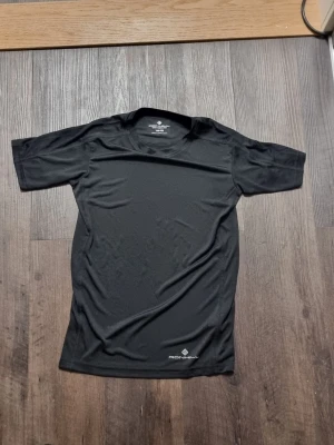Svart tränings-tshirt från Ronhill - INSTABOX!!! Snygg svart tränings-tshirt från Ronhill i funktionsmaterial som andas. T-shirten har rund halsringning, korta ärmar och en diskret logga nere vid fållen. Perfekt för löpning, gym eller annan träning.