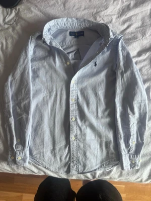 Ljusblå skjorta från Ralph Lauren - Klassisk ljusblå skjorta från Ralph Lauren med broderad logga på bröstet. Skjortan har knappar framtill och vid ärmsluten samt en snygg krage. Perfekt för en stilren och avslappnad look. De är ett äkta plagg. Funkar perfekt för 14 åringar. DM FÖR MER INFO!