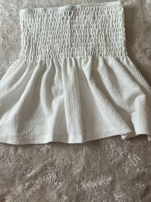 Vit bandeau tubtopp från Gina Tricot - Supersöt vit bandeau tubtopp från Gina Tricot i smockat tyg. Toppen är axelbandslös och har en stretchig, figurnära passform upptill med en lätt utställd nederdel. Perfekt till sommaren och enkel att styla med jeans eller kjol. Funkar som kjol eller topp . Pris går att diskutera .