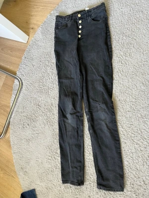 Svarta high waist skinny jeans - Säljer ett par svarta high waist skinny jeans med fem knappar framtill och klassiska fickor. Jeansen har bälteshällor och är i stretchigt denimtyg som sitter tight hela vägen ner. Perfekta för en trendig och bekväm look.