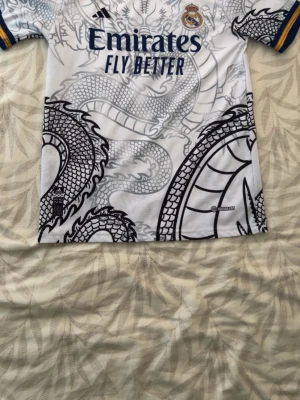 Real Madrid tröja Ronaldo nr 7 drake - Säljer en vit Real Madrid matchtröja från Adidas med stort svart drakmönster, blå detaljer och gula ränder på ärmarna. Tryck med 'Emirates Fly Better' på bröstet och 'Ronaldo 7' på ryggen. Perfekt för dig som älskar fotboll och vill sticka ut på planen. Den passar en man på 175-185 cm lång
