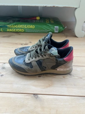 Valentino rockrunners camo sneakers med mockadetaljer - Snygga sneakers från Valentino med kamouflagemönster i grönt och grått, detaljer i mocka och läder samt en röd detalj på hälen. Skorna har gröna skosnören och låg profil. Perfekta för dig som vill sticka ut med exklusiva material och cool design.