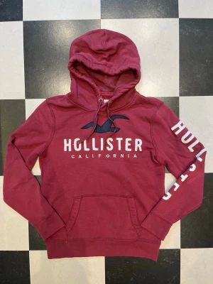 Hollister hoodie - Snygg rödhoodie från Hollister med stor logga och text på bröstet samt tryck på ena ärmen. Klassisk känguruficka framtill och justerbar huva med dragsko. Mjuk bomullsblandning som är skön att ha på sig.