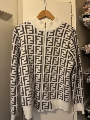 Tröja - Säljer en snygg ziptröja i vitt och brunt med det klassiska FF-mönstret över hela plagget. Tröjan har hög krage, dragkedja framtill och ribbade muddar. Perfekt för dig som gillar lyxiga streetwear-vibbar.