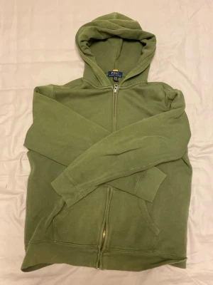 Olivgrön hoodie från Polo Ralph Lauren - Säljer en olivgrön hoodie från Polo Ralph Lauren i storlek xs. Hoodien har dragkedja framtill, två fickor och en klassisk huva. Tillverkad i mjukt bomullsmaterial som känns skönt mot huden. Perfekt för en avslappnad och stilren look.