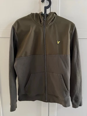 Olivgrön hoodie från Lyle & Scott - Snygg olivgrön hoodie med dragkedja från Lyle & Scott. Hoodien har tvådelad design med mörkare och ljusare partier, fickor framtill och den klassiska gula loggan på bröstet. Tillverkad i mjukt material och har ribbade muddar vid ärmar och nederkant.
