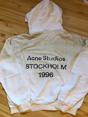 Vit hoodie Acne Studios Stockholm - Säljer en vit hoodie från Acne Studios med trycket 'Acne Studios STOCKHOLM 1996' på ryggen. Hoodien har klassisk huva och ribbade muddar vid ärmslut och nederkant. Perfekt för dig som gillar streetwear och enkel, stilren design.