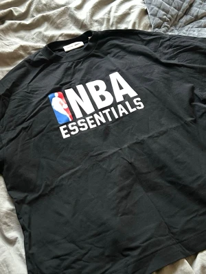 Svart NBA Essentials t-shirt - Snygg svart t-shirt från NBA Essentials med stor vit logga och NBA-symbol på bröstet. Klassisk passform och rund halsringning, perfekt för dig som gillar basket och streetwear.
