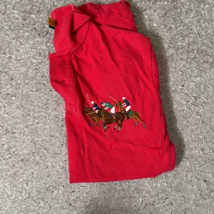 Röd pikétröja från Polo Ralph Lauren - Röd pikétröja från Polo Ralph Lauren med broderat polospel-motiv på bröstet och en stor blå siffra 3 på ärmen. Klassisk krage med knappar och korta ärmar. Perfekt för en sportig och stilren look.