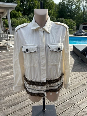 Vit jeansjacka med dekorativa detaljer - Stilren vit overshirt med axelklaffar, två bröstfickor med knapp och kontrastsömmar. Jackan har unika dekorativa band och fransar i brunt och vitt nedtill. Långärmad och tillverkad i ett mjukt bomullsmaterial.