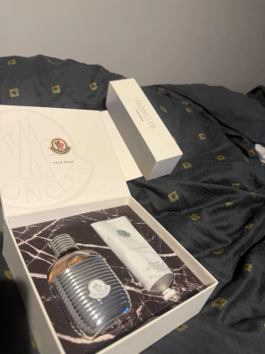 Moncler Pour Femme parfymset - Lyxigt parfymset från Moncler med en elegant parfymflaska i silver och glas samt en matchande vit tub. Flaskan har en unik ribbad design i metall och Moncler-logga framtill. Perfekt för dig som gillar exklusiva dofter och snygg design.