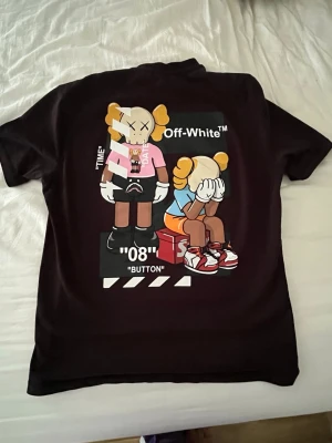 Svart Off-White t-shirt med tryck - Cool svart t-shirt från Off-White med stort färgglatt tryck på ryggen med KAWS-inspirerade figurer och Off-White-logga. Framsidan har en enkel vit Off-White-logga med två kryss. Klassisk passform och korta ärmar.