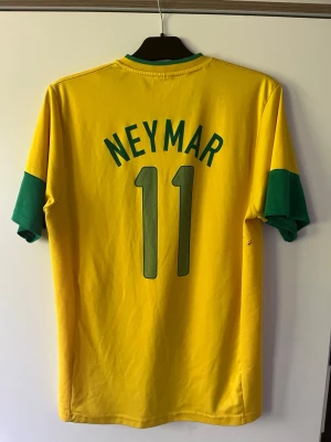 Brasilien Neymar 11 matchtröja Nike - Gul Brasilien fotbollströja från Nike med gröna detaljer, Neymar och nummer 11 på ryggen. Tröjan har officiellt CBF-märke och Nike-logga på bröstet samt gröna muddar på ärmarna. Tillverkad i lätt och ventilerande material, perfekt för match eller träning.