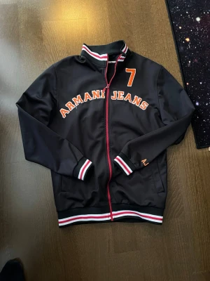 Svart jacka Armani Jeans - Svart jacka från Armani Jeans med röd dragkedja och vita, röda och gröna detaljer på krage, ärmslut och nederkant. Stor broderad text 'ARMANI JEANS' och siffran 7 på bröstet. Tillverkad i ett glansigt syntetmaterial och har sportig college-look.
