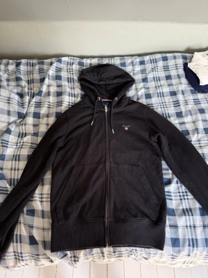 Svart hoodie med dragkedja från Gant - Säljer en svart hoodie från Gant med dragkedja framtill och två fickor. Klassisk huva med dragsko och Gant-logga broderad på bröstet. Tillverkad i mjukt bomullsmaterial som är skönt att ha på sig. Perfekt för en avslappnad och stilren look.