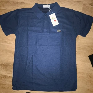 Lacoste Regular Fit Polo T-shirt- Marinblå (ny med taggar) storlek S - Helt ny Lacoste pikétröja i klassisk marinblå färg. Skick: Helt ny, aldrig använd. Passform: Regular Fit (bekväm, klassisk passform).  Material: Bomull.  Detaljer: Klassisk krage, två knappar framtill och den ikoniska broderade krokodilen på bröstet.  Färg: Marinblå. Mått: (S) Axelbredd: ca 40 cm .           Längd: ca 62 cm . Perfekt för både vardag och finare tillfällen. 