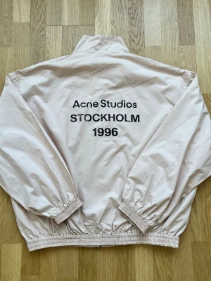 Beige vindjacka Acne Studios - Snygg beige vindjacka från Acne Studios med tryck 'STOCKHOLM 1996' på ryggen. Jackan har hög krage, resår i ärmslut och nederkant samt en avslappnad passform. Perfekt för dig som gillar minimalistisk och stilren design.