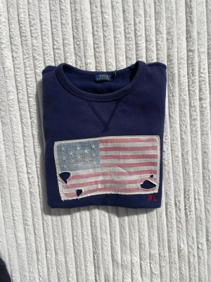 Mörkblå sweatshirt Polo Ralph Lauren - Mörkblå sweatshirt från Polo Ralph Lauren med amerikansk flagga i vintage-stil på bröstet. Flaggan har slitna detaljer och RL-broderi i rött. Tröjan har rund halsringning och ribbade muddar vid ärmar och nederkant. Perfekt för en avslappnad och cool look.