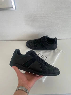 Svarta Gats från Maison Margiela - Säljer ett par svarta sneakers från Maison Margiela med klassisk low-top siluett. Skorna har en mix av mocka och läderpaneler, svarta skosnören och diskret logga på insidan. Perfekta för dig som gillar minimalistisk och stilren design.