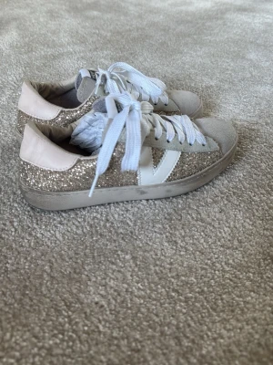 Glittriga sneakers från Victoria - Snygga sneakers från Victoria med guldglittriga sidor, vita detaljer och vita skosnören. Skorna har en låg profil och beige mockaliknande tåparti samt vit sula. Perfekta för dig som vill sticka ut med lite extra bling i vardagen.
