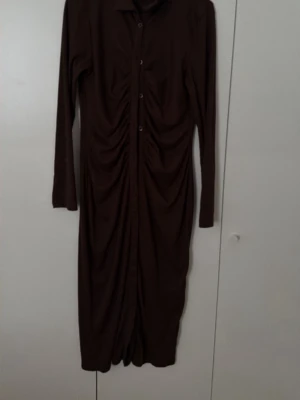 Brun långklänning med knappar - Snygg brun långklänning med långa ärmar och knappar framtill. Klänningen har rynkade detaljer längs hela framsidan som ger en cool och trendig look. Perfekt för dig som gillar stilrena plagg med en twist.