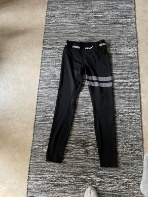 Svarta leggings från Stronger - Snygga svarta leggings från Stronger med logga i midjan och två gråa ränder på ena benet. Tight passform och hög midja, perfekta för träning eller chill. Materialet är stretchigt och följsamt.