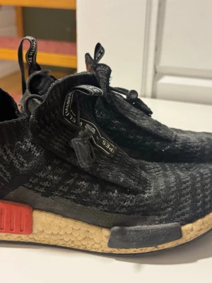 Adidas NMD City Sock svarta sneakers - Säljer ett par Adidas NMD City Sock sneakers i svart med stickad ovandel och elastisk mudd. Skorna har en beige Boost-sula med röda och svarta detaljer på sidan samt dragflikar med text på hälen och plösen. Perfekta för dig som gillar streetwear och bekväma skor.