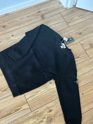 Svart sweatshirt från Stone Island - Svart sweatshirt från Stone Island med klassisk rund hals och ribbade muddar. Ikonisk logotyp-patch på vänster ärm. Tröjan är i mjukt material och har en stilren, enkel design som passar perfekt till streetwear.