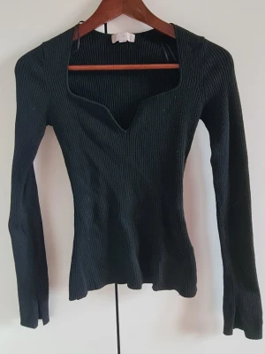 Svart ribbad långärmad topp - Snygg svart ribbad topp med lång ärm och unik v-ringad halsringning. Figurnära passform som framhäver siluetten och stretchigt material för extra komfort. Perfekt att styla med jeans eller kjol för en trendig look.