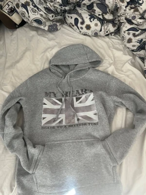 Grå hoodie med brittisk flagga - Snygg grå hoodie med stor ficka framtill och tryck av brittiska flaggan på bröstet. Texten 'MY HEART BEATS TO A BRITISH TUNE' står under flaggan. Mjuk bomullsmix och klassisk huva med dragsko. Perfekt för dig som gillar streetstyle och coola prints.