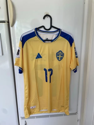 Svensk landslagströja Adidas gul/blå - Säljer en officiell svensk landslagströja från Adidas i gul med blå detaljer och nummer 17 på framsidan. Tröjan har korta ärmar, broderad landslagssköld och Adidas-logga. Tillverkad i lätt och ventilerande material, perfekt för fotboll eller träning.