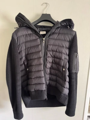 Moncler cardigan - Snygg svart dunjacka från Moncler med stickade ärmar och huva. Jackan har quiltad front, två fickor med tryckknappar och en ärmficka med dragkedja. Klassisk Moncler-logga på ärmen och ribbade muddar för extra komfort. NFC tagg fungerar den är använd en fåtal gånger men är i riktigt bra skick. Storleken är L vilket motsvarar ungefär S till M