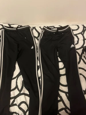 Svarta Adidas mjukisbyxor med vita ränder - Säljer ett par klassiska svarta Adidas mjukisbyxor med vita ränder längs benen och Adidas-logga på låret. Byxorna har resår i midjan och är tillverkade i ett mjukt och bekvämt syntetmaterial. Perfekta för chill eller träning!säljer en för 89 två 149