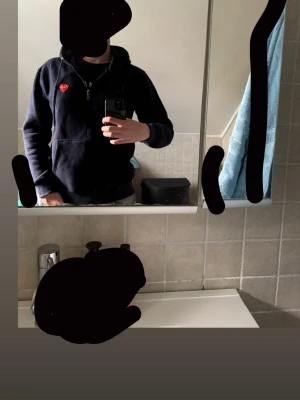 Marinblå hoodie med ett hjärta  - Säljer en marin blå hoodie med dragkedja och huva. Klassisk röd hjärtlogga med ögon på bröstet. fickor framtill och ribbade muddar. Perfekt för en clean och stilren look. Mer som en L så lite liten i strl! Lappen på tröjan är lite utnött men inget som märks vid användning annars bra!