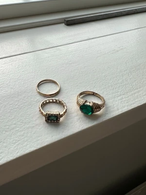 Tre guldringar med gröna stenar - Säljer tre snygga ringar i guldton. En enkel tunn ring, en med rektangulär grön sten och pärldetaljer runt, samt en med stor hjärtformad grön sten. Perfekta för att mixa och matcha till olika outfits.