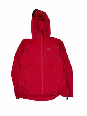 Röd vindjacka från Arc'teryx med huva - Snygg röd vindjacka från Arc'teryx med hel dragkedja, justerbar huva och flera praktiska fickor med dragkedja. Jackan har en sportig design och är tillverkad i lätt, slitstarkt material som passar perfekt för utomhusaktiviteter. Givetvis äkta!