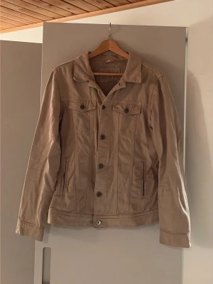 Oveshirt Beige jeansjacka  - Stilren beige jeansjacka med raka linjer och klassisk krage. Jackan har två bröstfickor med lock och knapp samt två sidofickor. Knappstängning framtill och diskreta sömdetaljer ger en clean och tidlös vibe.