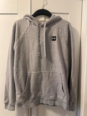 Grå hoodie från Under Armour - Snygg grå hoodie från Under Armour med klassisk känguruficka och justerbar dragsko i huvan. Tröjan har ribbade muddar vid ärmslut och nederkant samt en liten logga på bröstet. Perfekt för en avslappnad och sportig stil.