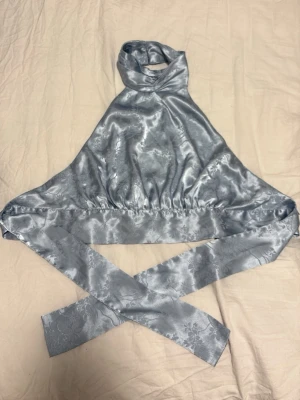 Silvrig halterneck topp i satin - Trendig silvrig halterneck topp i glansigt satinliknande material. Toppen har breda knytband i midjan och runt halsen för en snygg omlott-effekt. Perfekt för dig som vill sticka ut med en unik och elegant look.
