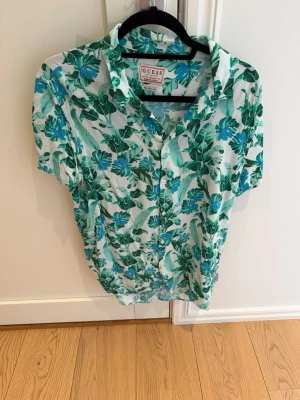 GUESS Tropical Print Shirt – Regular Fit, Strl S – Kanonskick - Stilren och somrig skjorta från GUESS med ett snyggt grönt botaniskt mönster som ger en avslappnad men samtidigt trendig känsla. Perfekt för semestern, sommarens tillställningar eller för dig som vill addera lite tropisk elegans till garderoben.  ✔ Märke: GUESS ✔ Storlek: S ✔ Passform: Regular Fit ✔ Grönt botaniskt/tropiskt mönster ✔ Bekväm och luftig modell ✔ Kanonskick  Skjortan är mycket välvårdad och kommer från ett djur- och rökfritt hem. 