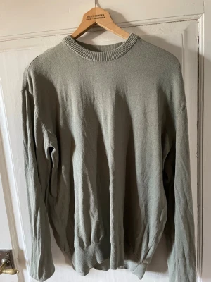 Ljusgrå basic tröja med rund hals - En enkel ljusgrå tröja med rund halsringning och långa ärmar. Tröjan har ribbade muddar vid hals, ärmslut och nederkant. Perfekt för en clean och avslappnad stil. Materialet ser ut att vara mjukt och bekvämt, troligen bomull eller bomullsblandning.