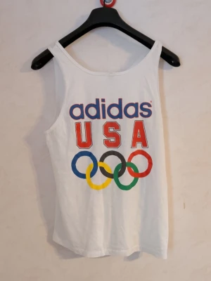 Vit Adidas USA linnetopp vintage 80-tal - vitt linne från Adidas med stort tryck framtill: 'adidas USA' och de klassiska OS-ringarna i blått, gult, svart, grönt och rött. Perfekt för dig som gillar sportig stil och vill sticka ut med en retrovibe. Tunn och skön bomull som passar till sommaren.