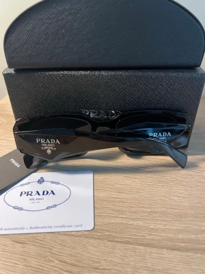 Svarta solglasögon från Prada - Stilrena svarta solglasögon från Prada med rektangulära bågar och diskret logga på skalmen. Kommer med originaletui och äkthetskort. Perfekta för dig som vill ha en klassisk och lyxig accessoar till sommaren.