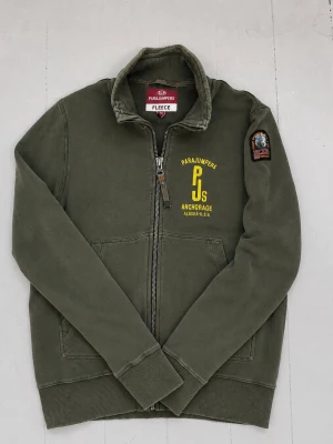 Olivgrön fleecejacka Parajumpers S - Olivgrön fleecejacka från Parajumpers med dragkedja framtill, två fickor och gul logga på bröstet. Hög krage och ribbade muddar. Patch på ena ärmen och tryck med texten 'Anchorage Alaska-USA'. Perfekt för lager-på-lager och streetwear.