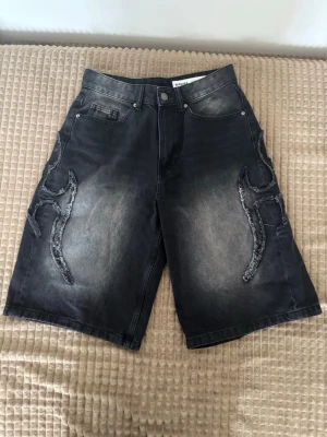 Svarta denimshorts  - Säljer ett par svarta denimshorts med grå tvätt och unika broderade detaljer på sidorna. Shortsen har fem fickor, knappgylf och en loose passform som ger en avslappnad vibe. Perfekta för dig som vill sticka ut med coola detaljer.